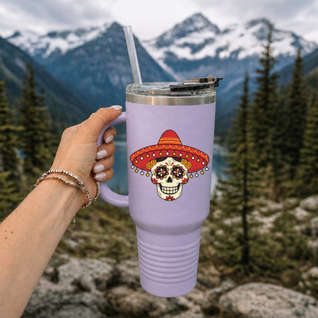 A hand holds a lavender insulated tumbler with a clear straw in front of a mountain and forest background. The tumbler features a sugar skull graphic wearing a red and orange sombrero.