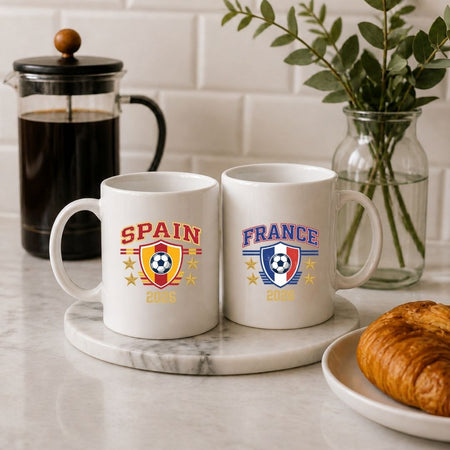 A close up of two white ceramic mugs sitting on a marble coaster next to a coffee press and pastry, printed with vintage Spain and France soccer tournament crests.