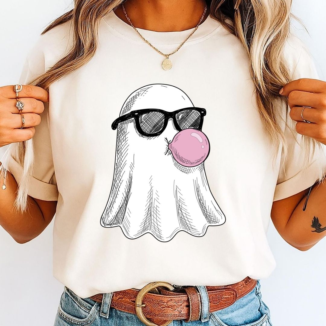 A woman wearing a cream-colored t-shirt with a graphic of a ghost wearing black sunglasses and blowing a pink bubblegum bubble. The image is cropped from the chest down, showing the t-shirt, blue jeans, a brown leather belt, and the model's hands.