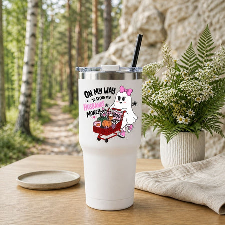 A white stainless steel travel tumbler with a black straw, featuring the ghost shopping Halloween design, set outdoors with a forest background.