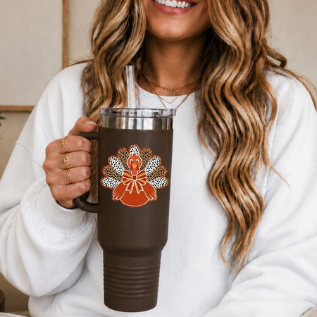 A woman in a white sweatshirt holds a dark brown insulated tumbler with a clear lid and straw. The tumbler features a decorative orange and white turkey design with rhinestone-like detailing on the front.