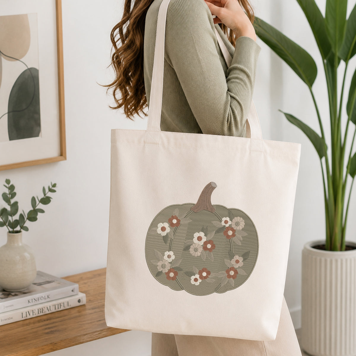 A woman wearing a sage green ribbed sweater carries a cream canvas tote bag with a printed pumpkin design featuring white, pink, and red flowers on a muted green background. The scene is set in a bright, modern room with a wooden table, books, a framed print, and a potted plant.