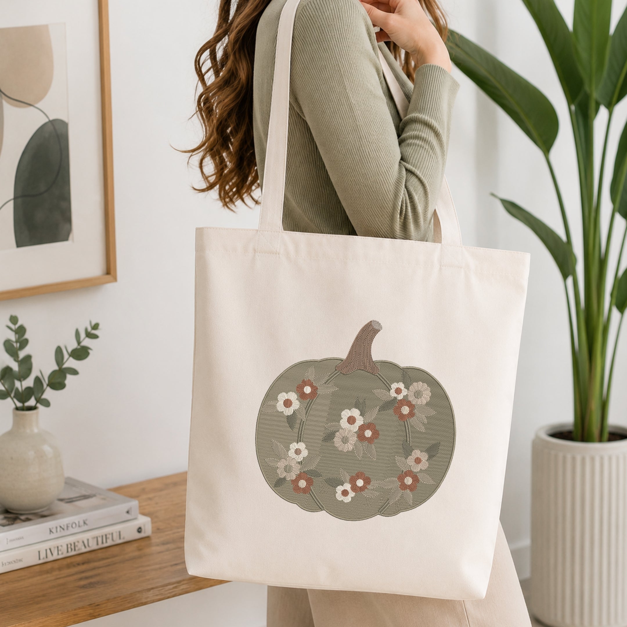 A woman wearing a sage green ribbed sweater carries a cream canvas tote bag with a printed pumpkin design featuring white, pink, and red flowers on a muted green background. The scene is set in a bright, modern room with a wooden table, books, a framed print, and a potted plant.