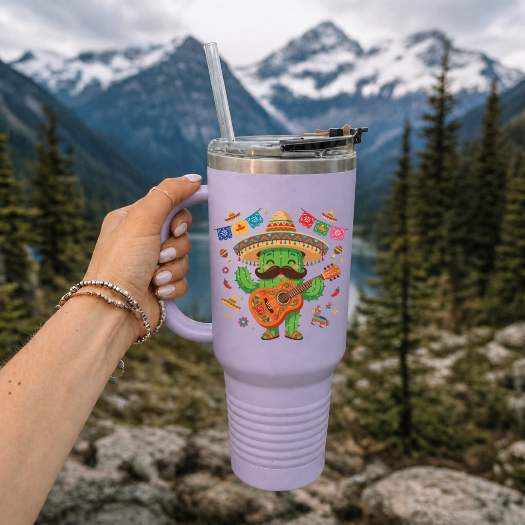 A lavender insulated tumbler with a clear lid and straw, held by a hand against a mountain and forest background. The tumbler features a colorful graphic of a cactus wearing a sombrero and playing a guitar.
