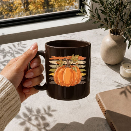 A hand holds a dark brown ceramic mug featuring a graphic of an orange pumpkin with autumn leaves and a sunset background. The scene is set on a light stone tabletop near a window with a potted plant and a candle in the background.