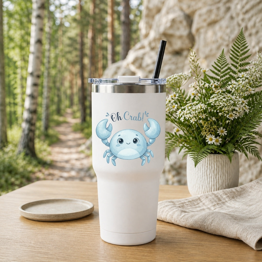 A white stainless steel tumbler with a lid and straw, decorated with a blue cartoon crab graphic and the text "Oh Crab!", sitting on a wooden table outdoors.