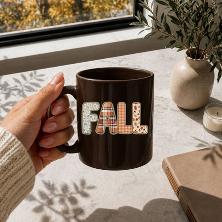 A hand holds a dark brown ceramic mug with the word 'FALL' spelled out in decorative patchwork letters, set on a light stone surface near a window with a plant and candle in a cozy indoor setting.