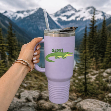 A hand holding a large lavender insulated travel tumbler with a handle and straw, featuring an alligator graphic against a scenic mountain and lake background.