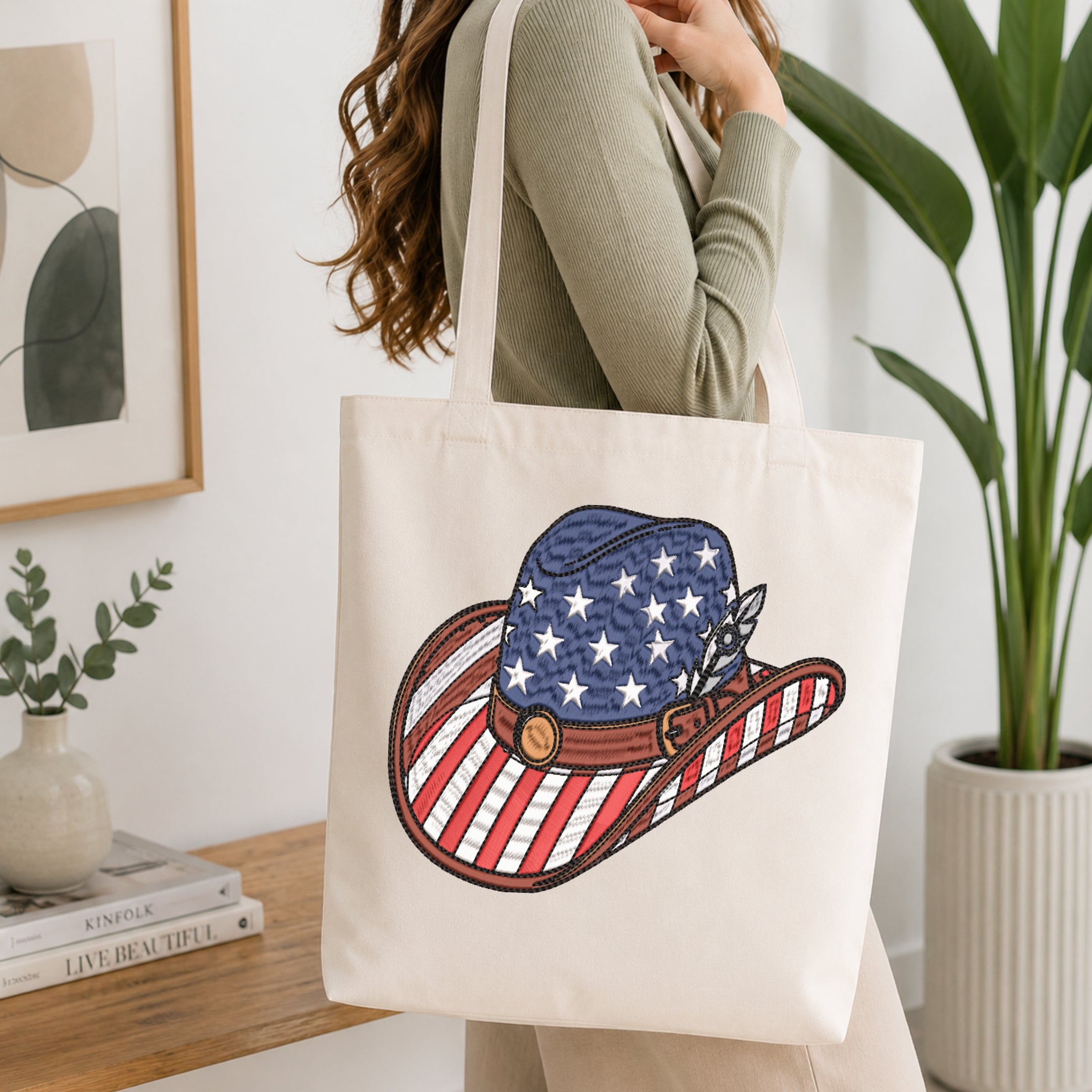 A woman wearing a sage green ribbed sweater carries a cream canvas tote bag with a large embroidered cowboy hat design featuring an American flag pattern. The setting is a bright, modern room with a wooden shelf, framed art, and a potted plant.