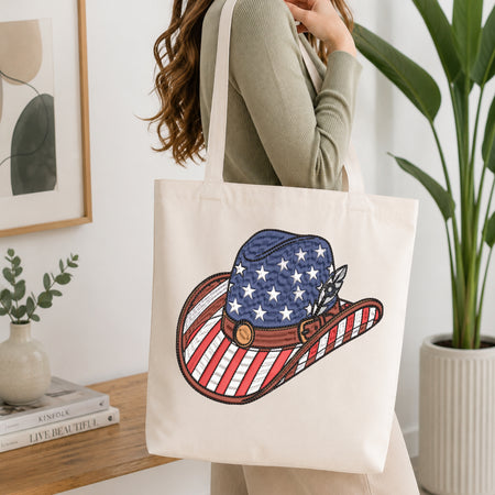 A woman wearing a sage green ribbed sweater carries a cream canvas tote bag with a large embroidered cowboy hat design featuring an American flag pattern. The setting is a bright, modern room with a wooden shelf, framed art, and a potted plant.