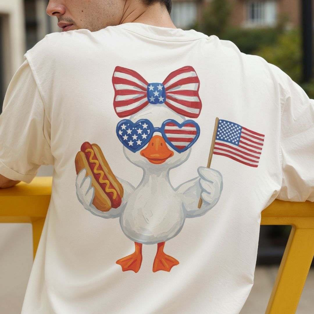 Person wearing a white t-shirt with a cartoon duck holding an American flag and a hot dog, with a blurred outdoor background.