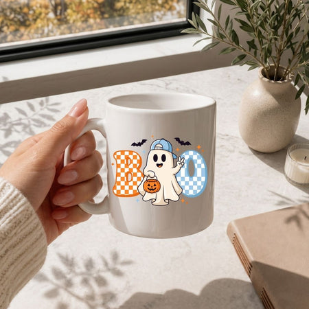 Hand holding a white coffee mug printed with a cute Halloween ghost graphic and checkered "BOO" letters, resting on a marble surface.