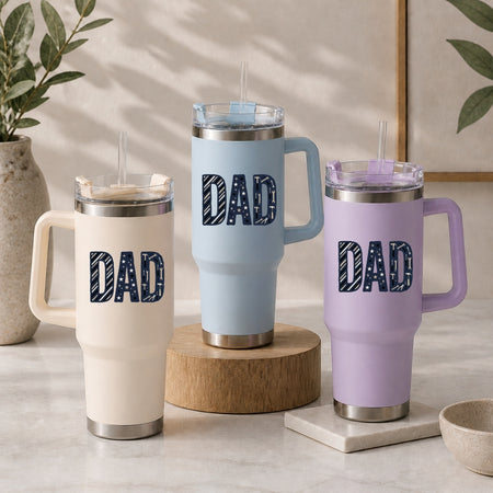 Three insulated handled tumblers with clear lids and straws are arranged on a light stone surface. Each tumbler has a large 'DAD' graphic on the front in a dark patterned fill.
