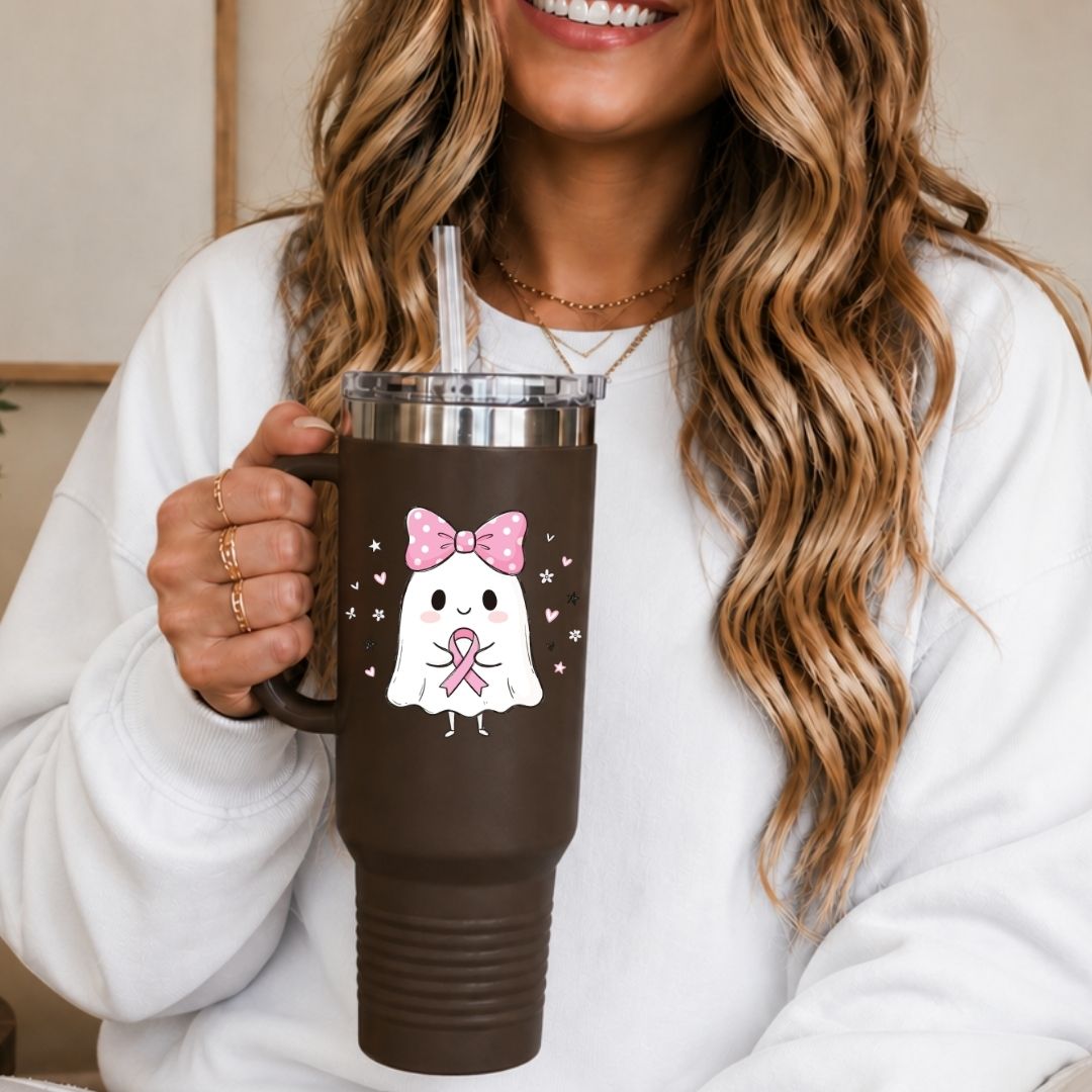 A woman in a white sweatshirt holds a dark brown insulated tumbler with a clear lid and straw. The tumbler features a cute cartoon ghost wearing a pink bow and pink ribbon, surrounded by small pink hearts.