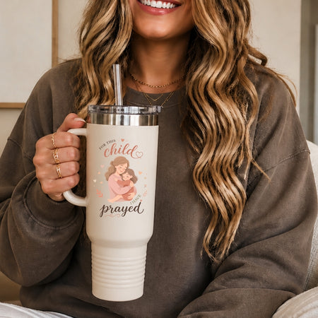 A person holding a cream-colored stainless steel tumbler with a straw, decorated with a mother-and-child illustration and the text "For this child I have prayed."