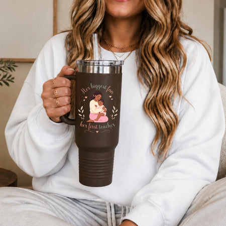 A woman holding a dark brown insulated travel tumbler with a handle, featuring the sweet illustration of a mother and child with text reading Her biggest fan, her first teacher.