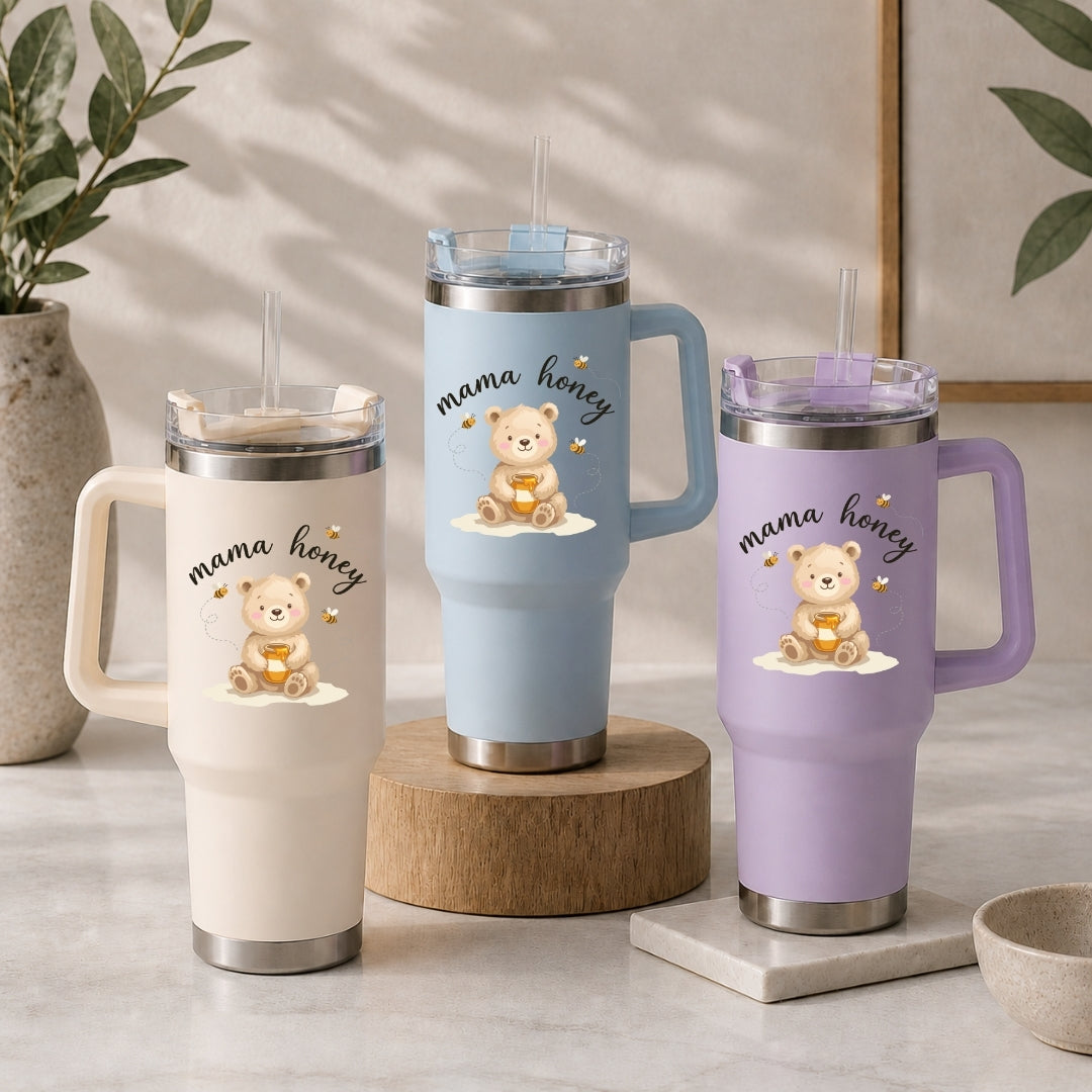 Three handled insulated tumblers with clear lids and straws are displayed on a light stone surface. Each tumbler has a different pastel body color: cream, light blue, and lavender, with a matching bear illustration and the words 'mama honey' printed on the front.