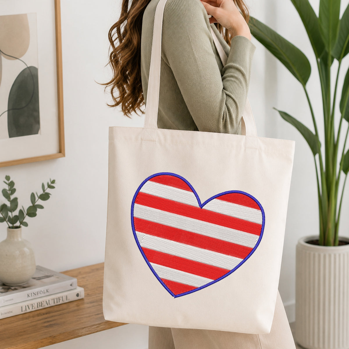 A woman wearing a sage green ribbed sweater carries a cream canvas tote bag with a large red and white striped heart outlined in blue on the front, set in a bright, modern room with a wooden table and potted plants.