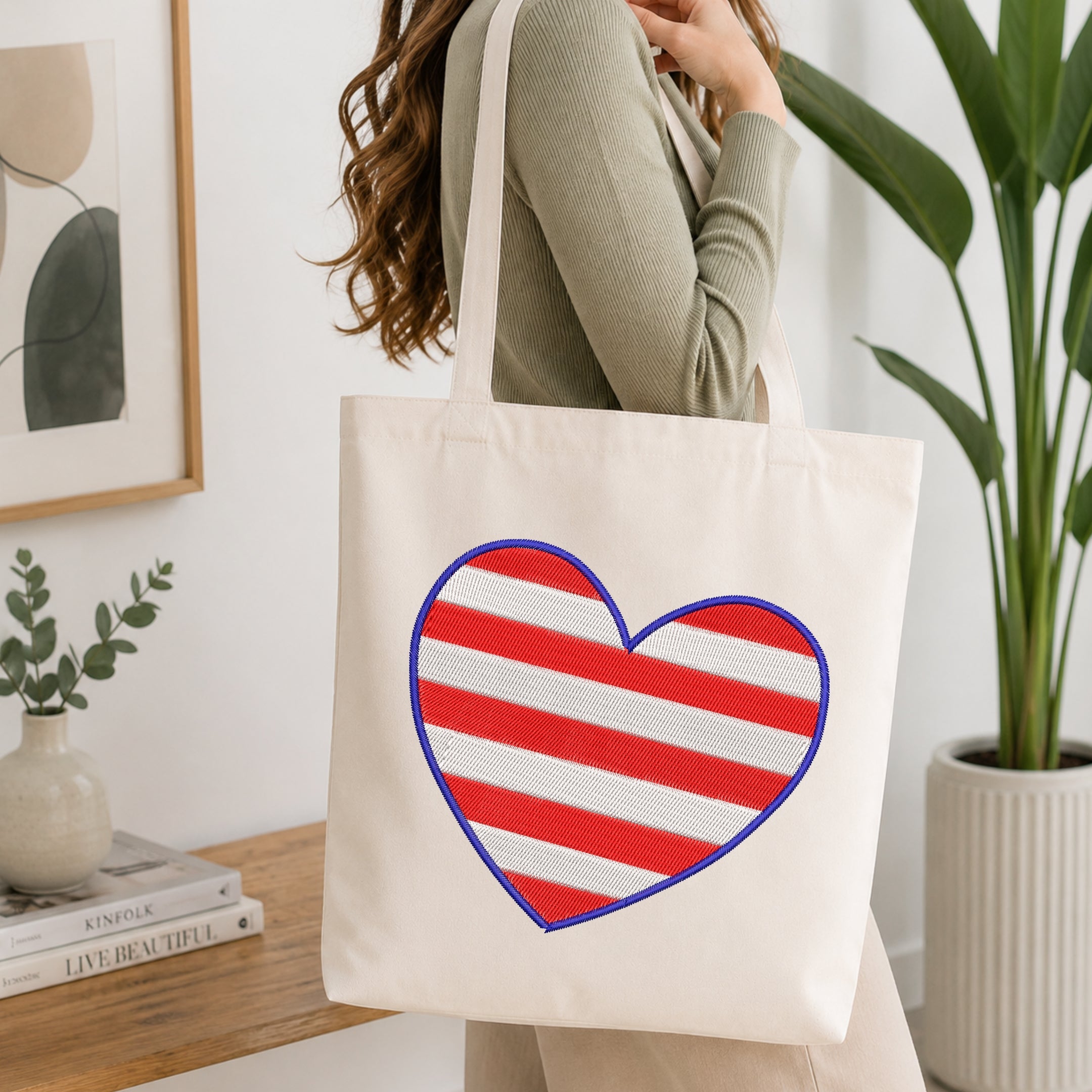 A woman wearing a sage green ribbed sweater carries a cream canvas tote bag with a large red and white striped heart outlined in blue on the front, set in a bright, modern room with a wooden table and potted plants.