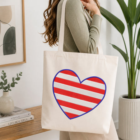 A woman wearing a sage green ribbed sweater carries a cream canvas tote bag with a large red and white striped heart outlined in blue on the front, set in a bright, modern room with a wooden table and potted plants.
