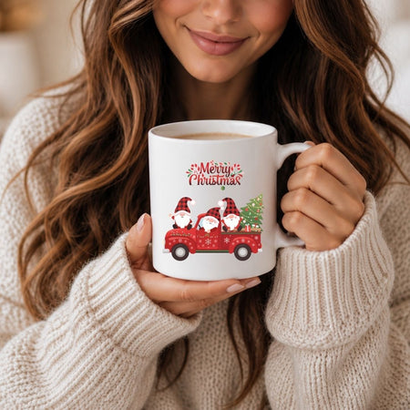 A woman in a cream knit sweater holds a white ceramic mug with a red truck carrying three gnomes and the words 'Merry Christmas' printed on the front.