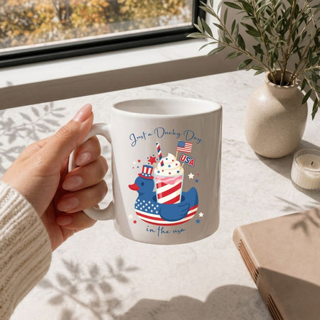 A hand holding a clean white ceramic coffee cup decorated with the red, white, and blue patriotic drink cup design on a bright marble table.