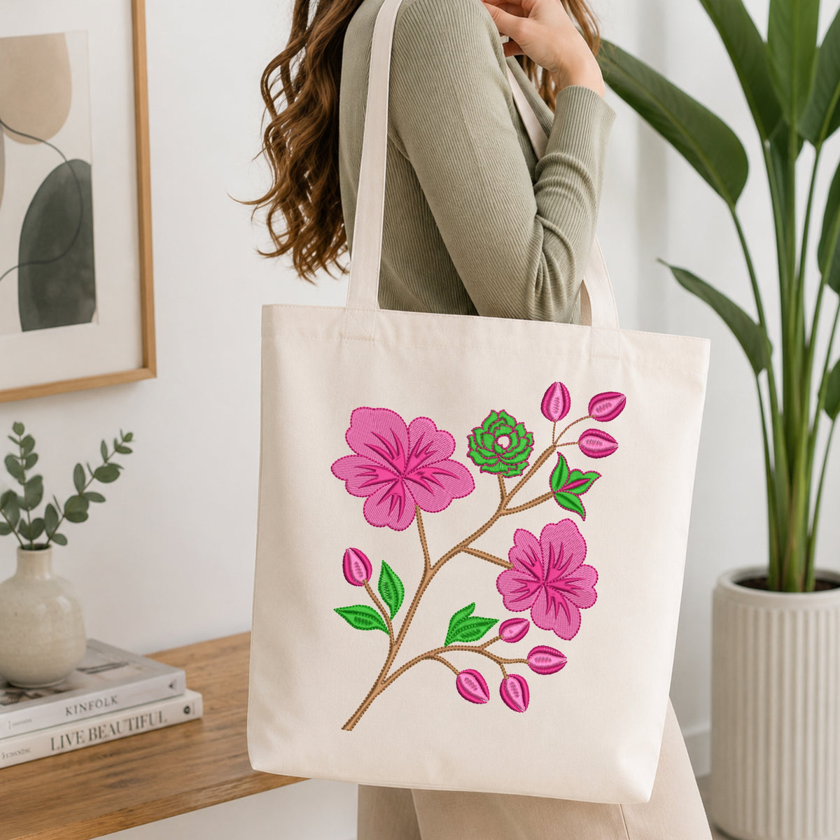 A woman wearing a sage green ribbed sweater carries a cream canvas tote bag with a colorful embroidered pink flower branch design, set in a bright, modern room with a wooden table and potted plants.