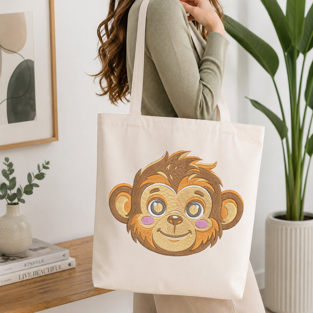A person wearing a sage green ribbed long-sleeve top carries a cream canvas tote bag featuring a large embroidered cartoon monkey face on the front, set in a bright, modern room with a wooden table, books, and a potted plant.