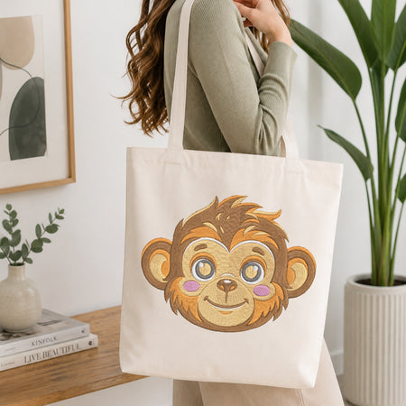 A person wearing a sage green ribbed long-sleeve top carries a cream canvas tote bag featuring a large embroidered cartoon monkey face on the front, set in a bright, modern room with a wooden table, books, and a potted plant.