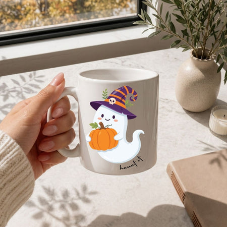 A hand holds a white ceramic mug featuring a cute cartoon ghost wearing a purple witch hat and holding an orange pumpkin. The mug is set on a light marble tabletop near a window, with a small potted plant, candle, and book in the background.