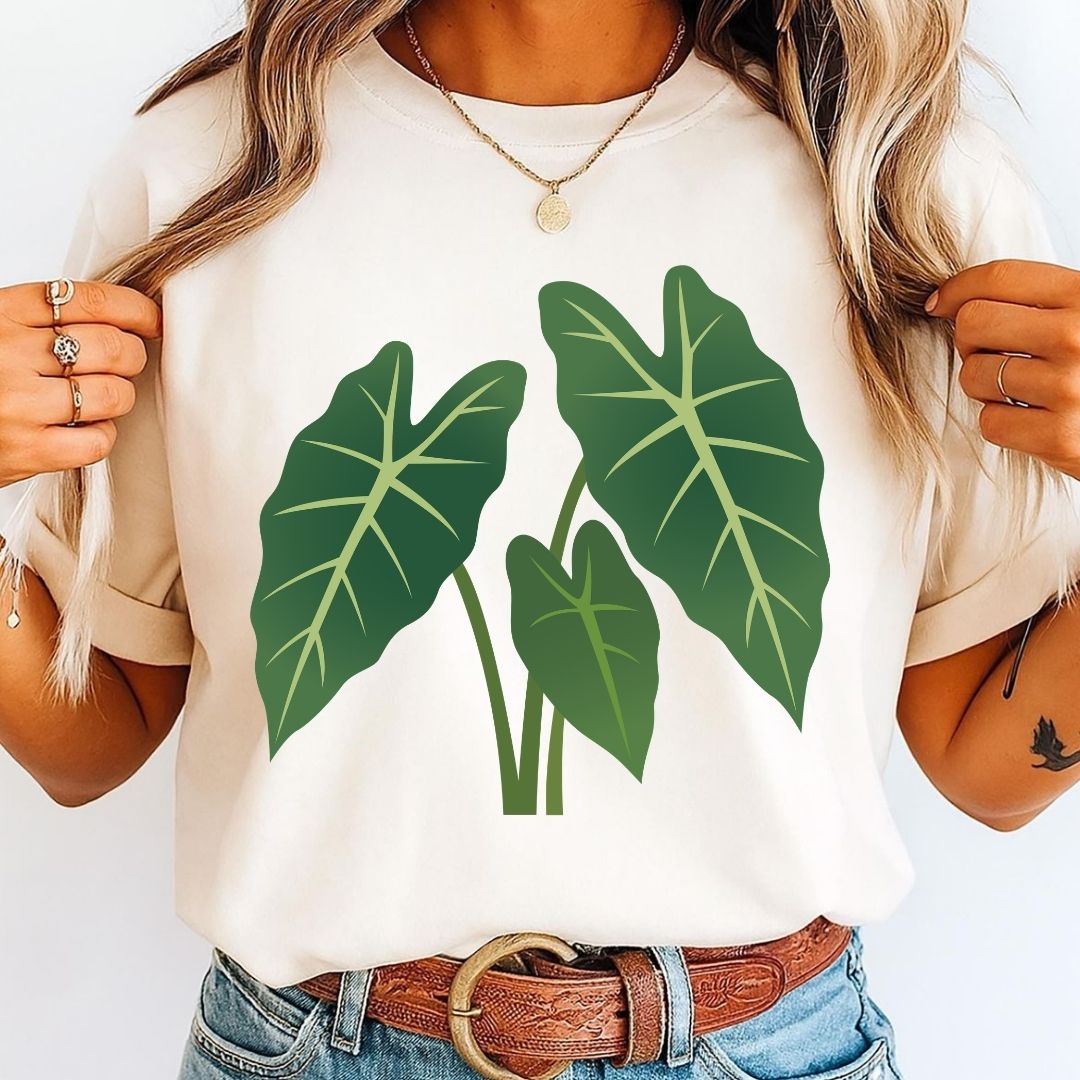 A woman wearing a cream-colored short-sleeve t-shirt with a green tropical leaf graphic on the front, paired with blue jeans and a brown leather belt.
