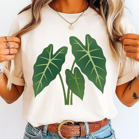 A woman wearing a cream-colored short-sleeve t-shirt with a green tropical leaf graphic on the front, paired with blue jeans and a brown leather belt.