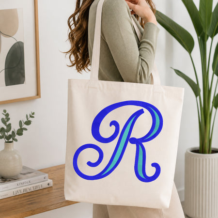 A woman wearing a sage green ribbed sweater carries a cream canvas tote bag with a large blue and teal monogram letter 'R' printed on the front, set in a bright, modern room with a wooden console table and a potted plant.