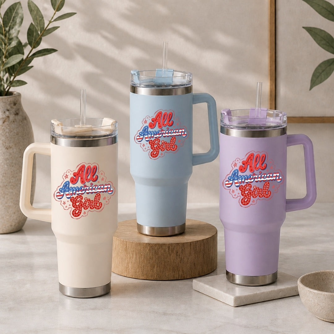 Three insulated handled tumblers with clear lids and straws are displayed on a light stone surface. The tumblers are in cream, light blue, and lavender, each printed with the phrase "All American Girl" in red, white, and blue script.