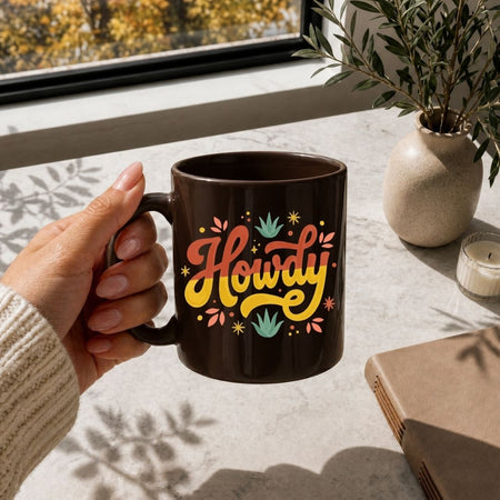 A hand with manicured nails holding a glossy brown ceramic coffee cup decorated with the groovy "Howdy" graphic next to a leather book.