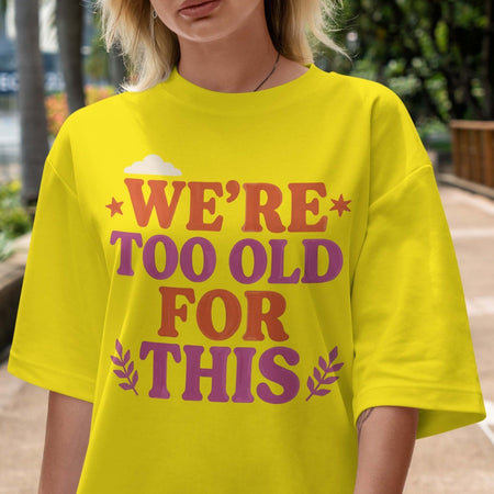 Person wearing a yellow t-shirt with text 'WE'RE TOO OLD FOR THIS' in an outdoor setting.