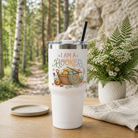 A white insulated tumbler with a clear lid and black straw sits on a wooden table in a forest setting. The tumbler has a printed design that reads “I AM A HOOKER” above a basket, fishing rod, reel, and bait illustration.