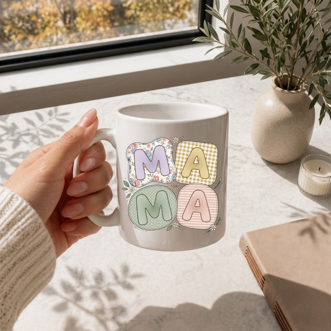 Hand holding a white ceramic coffee mug printed with the adorable quilted "MAMA" graphic and small daisy accents.