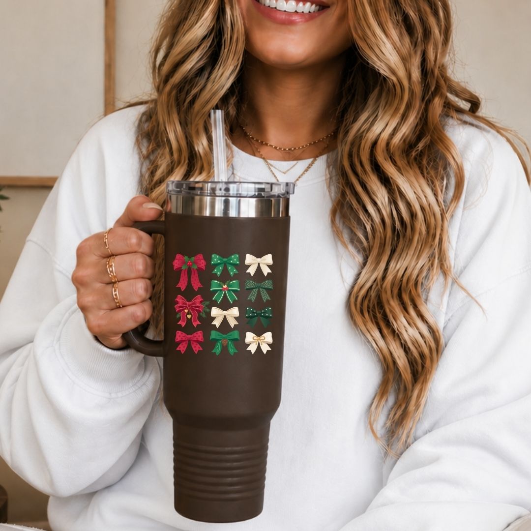 A woman in a white sweatshirt holds a brown insulated tumbler with a clear lid and straw. The tumbler is decorated with a grid of small gift bows in red, green, and cream, with the word 'Merry' visible in one design.