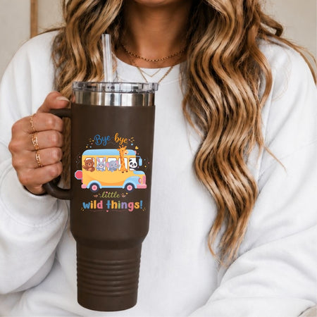 A person holding a brown stainless steel tumbler with a straw, decorated with a school bus graphic featuring cartoon animals and the text "Bye-bye little wild things!"