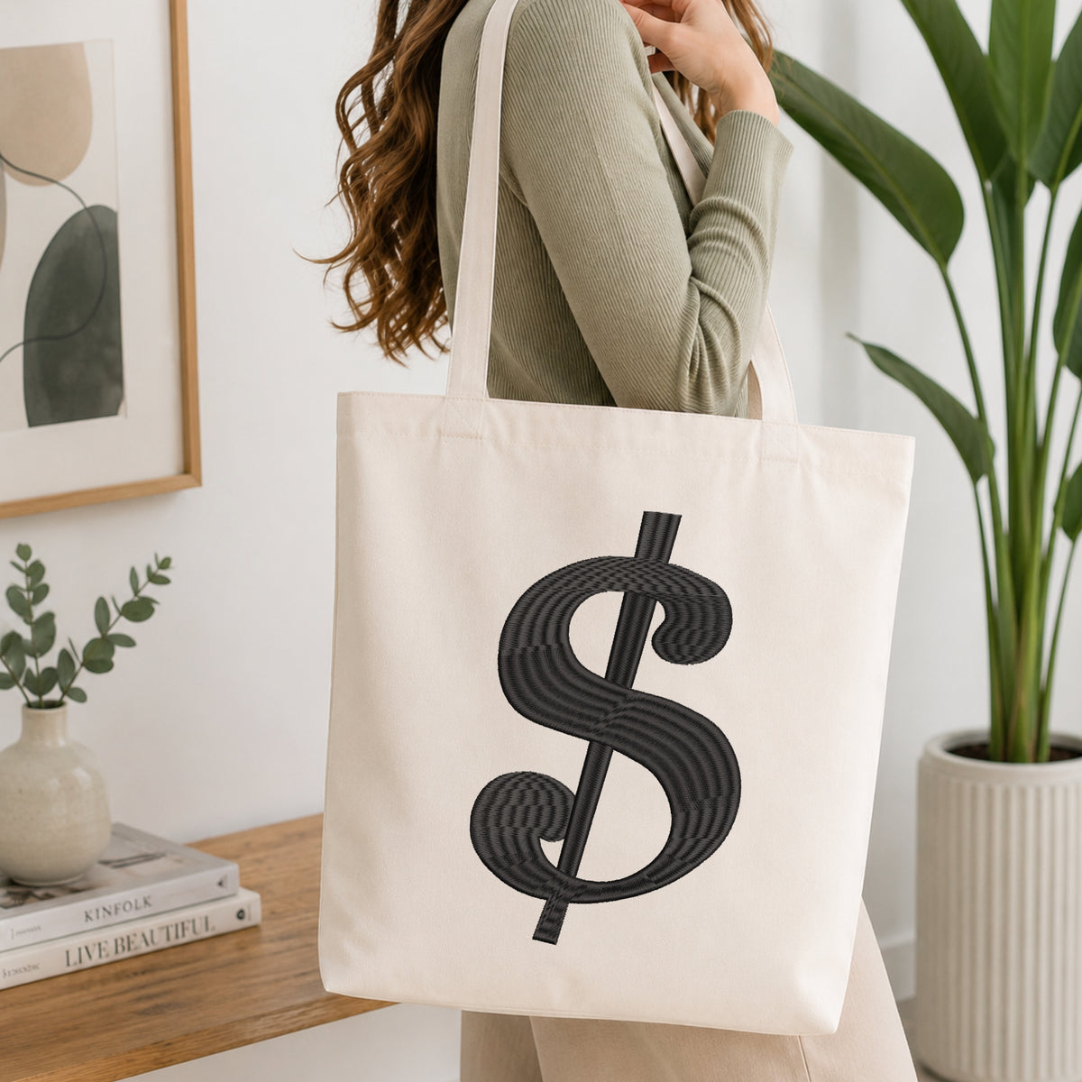A woman wearing a sage green ribbed top carries a cream canvas tote bag with a large black dollar sign graphic printed on the front, set in a bright, modern room with a wooden table, books, and a potted plant.