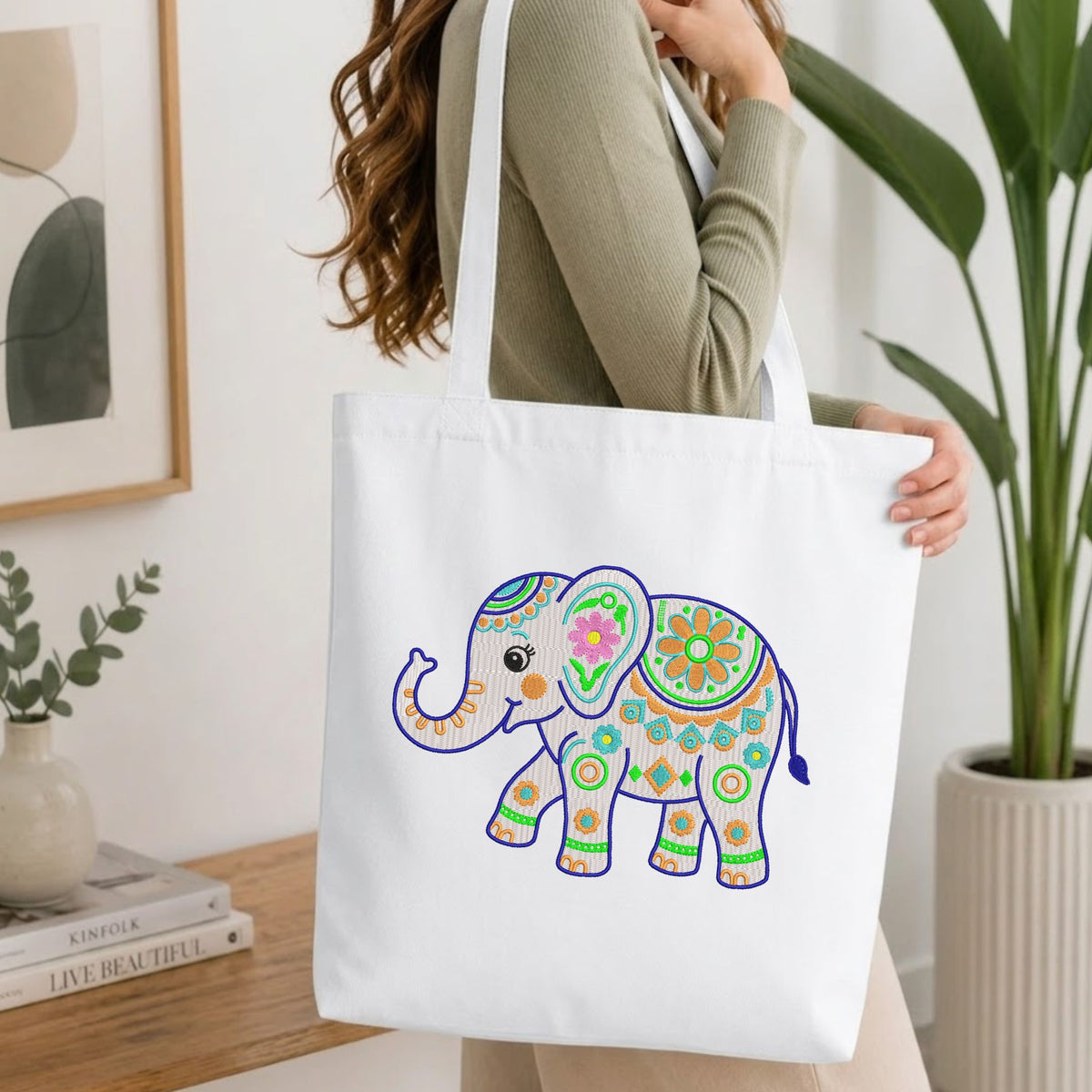 Woman carrying a white natural canvas tote bag featuring an embroidered folk art elephant design in a bright, modern interior setting.