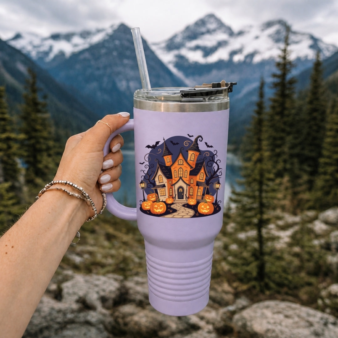 A hand holds a lavender purple tumbler with a clear straw in front of a mountain and forest background. The tumbler features a graphic of a haunted house surrounded by pumpkins.