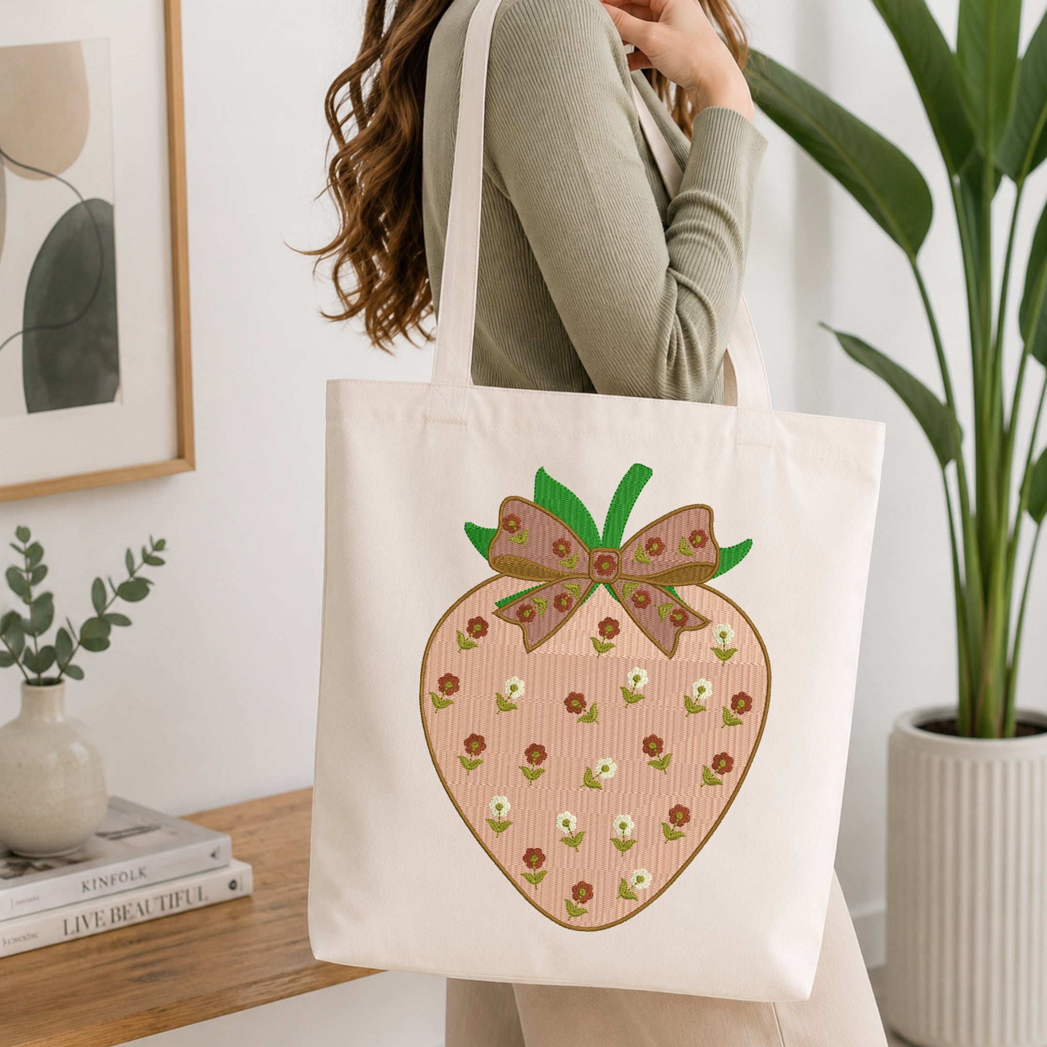 A woman carrying a canvas tote bag embroidered with a light pink strawberry featuring a floral pattern and a bow in a brightly lit room.