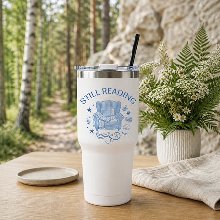A white insulated travel tumbler with a handle and black straw, featuring a custom blue "Still Reading" bookish design, placed on a outdoor table with a forest trail background.