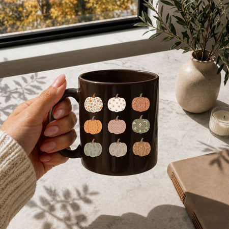 A hand holds a dark brown ceramic mug decorated with a grid of small autumn pumpkins in various colors and patterns, set on a light stone tabletop near a window with a plant and candle in the background.