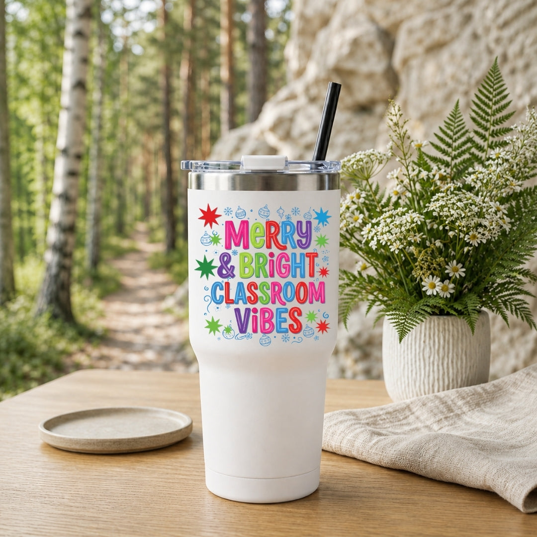 A white insulated tumbler with a clear lid and black straw sits on a wooden table in a forest setting. The front of the tumbler reads “Merry & Bright Classroom Vibes” in colorful lettering with small star and swirl accents.