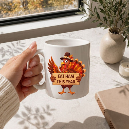A hand holds a white ceramic mug featuring a cartoon turkey wearing a pilgrim hat and holding a sign that reads 'EAT HAM THIS YEAR.' The mug is photographed in a bright indoor setting near a window, with a vase, candle, and book visible in the background.