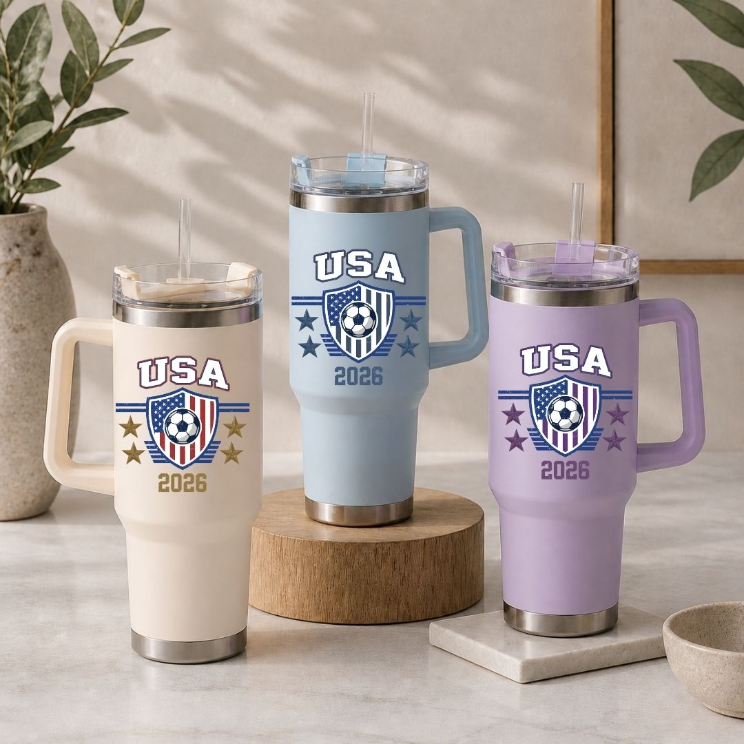 Three insulated tumblers in beige, light blue, and purple, each featuring the retro USA 2026 soccer shield design.