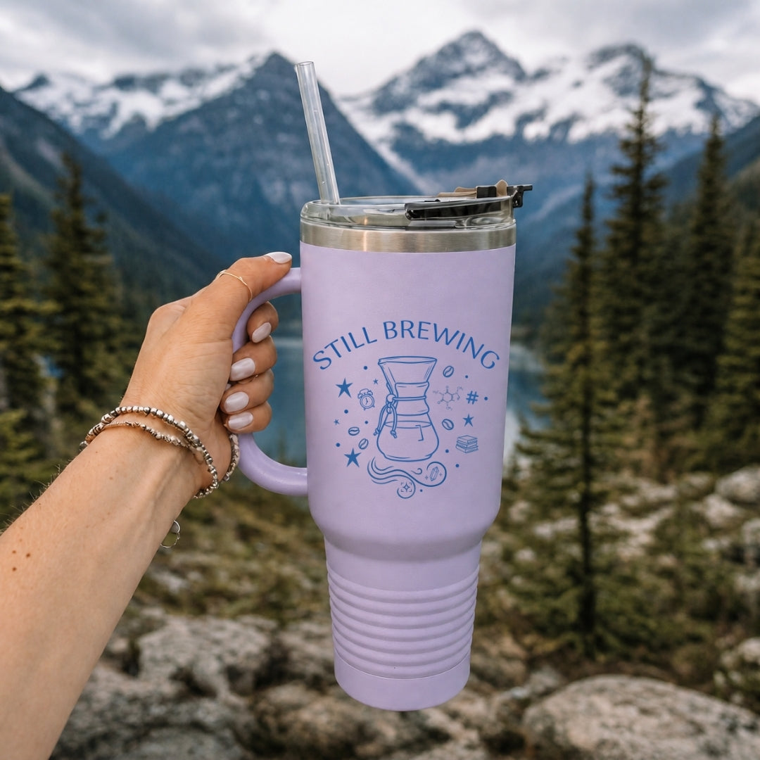 Hand holding a pastel lavender purple insulated travel tumbler with a handle and straw, featuring a custom blue "Still Brewing" coffee illustration against a scenic mountain lake background.